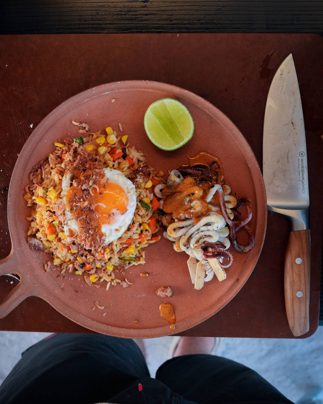 Overhead view of a rustic clay plate with a serving of nasi goreng topped with a runny fried egg, a side of grilled squid skewers drizzled with sauce, and a halved lime. A chef’s knife rests beside the dish, and a person’s legs are visible in the frame below.